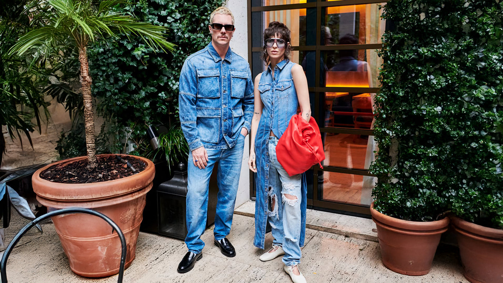 A stylish duo rocks bold double denim: he in a utility jacket, jeans, and loafers; she in a sleeveless vest over ripped jeans with a red bag.