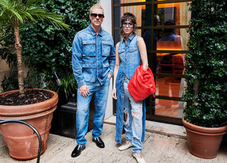 A stylish duo rocks bold double denim: he in a utility jacket, jeans, and loafers; she in a sleeveless vest over ripped jeans with a red bag.