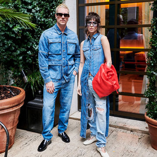 A stylish duo rocks bold double denim: he in a utility jacket, jeans, and loafers; she in a sleeveless vest over ripped jeans with a red bag.