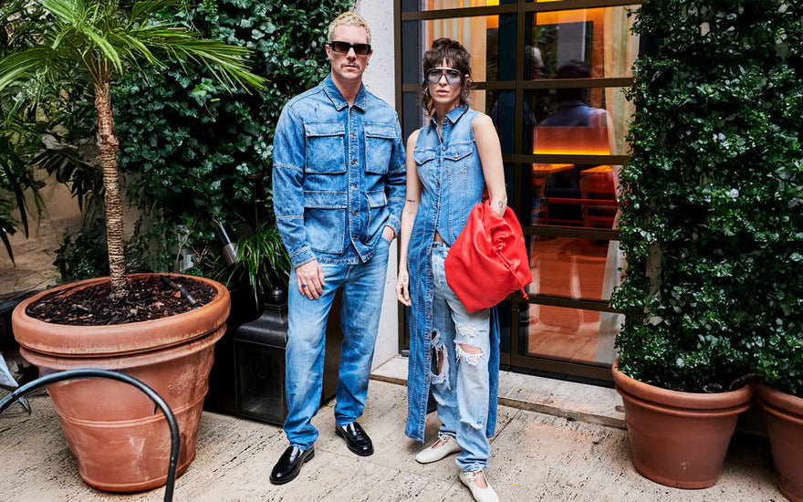 A stylish duo rocks bold double denim: he in a utility jacket, jeans, and loafers; she in a sleeveless vest over ripped jeans with a red bag.